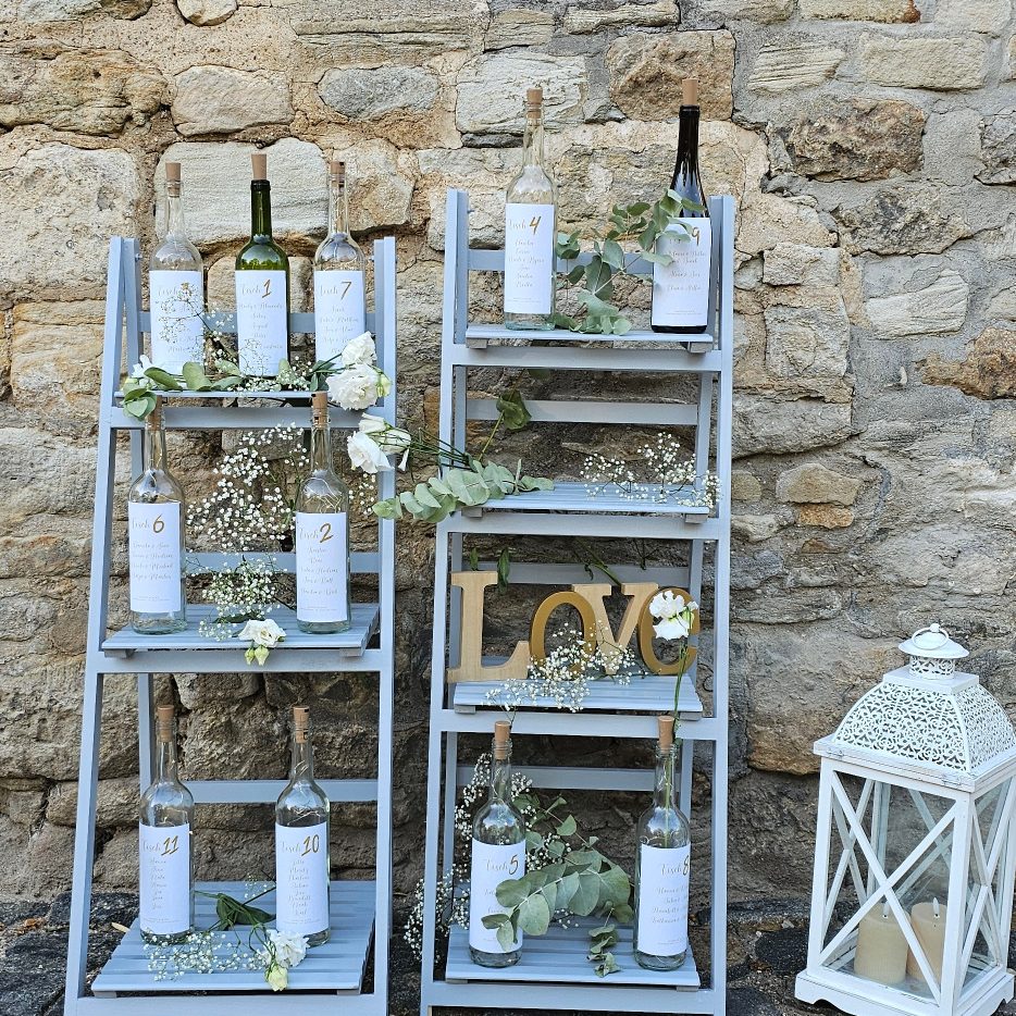 Regal mit Weinflaschen, Blumen und einem "Love"-Schild vor einer steinernen Wand.