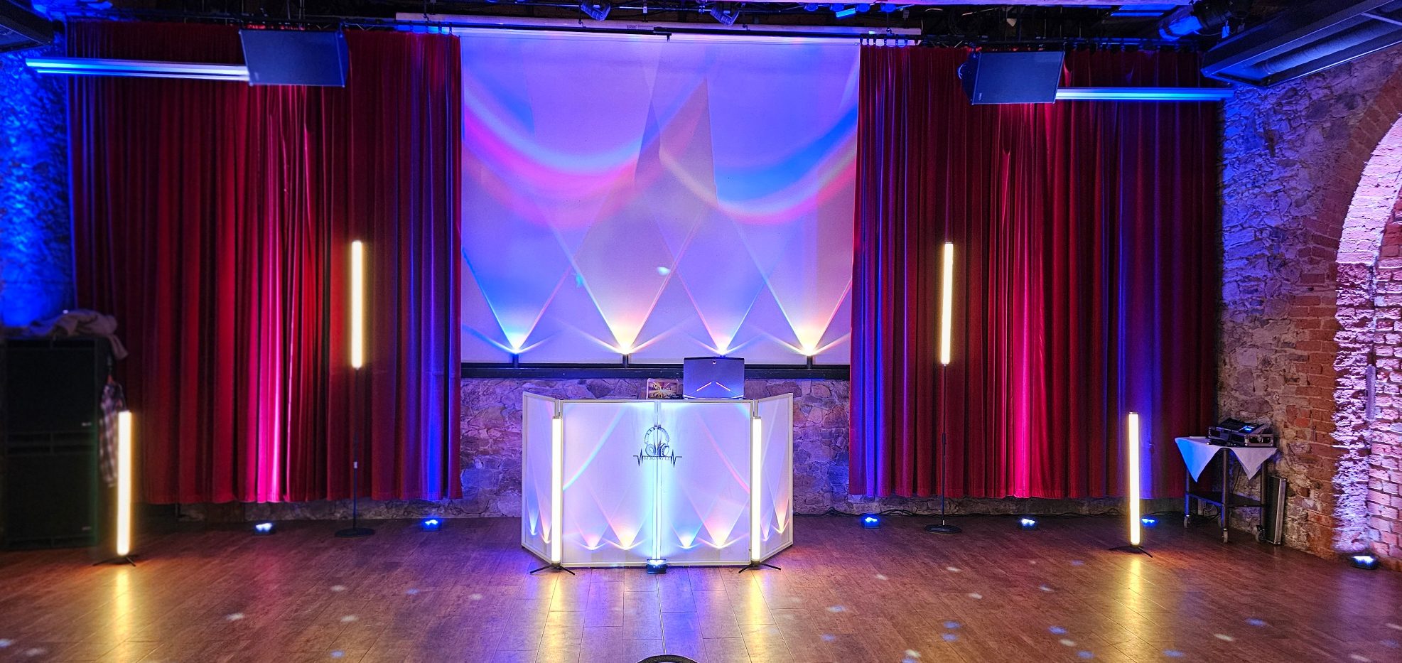 Kloster Nimbschen 7 Ein festlich dekorierter Veranstaltungsort mit rotem Samtvorhang und bunten Uplights sowie einem beleuchteten DJ Pult.